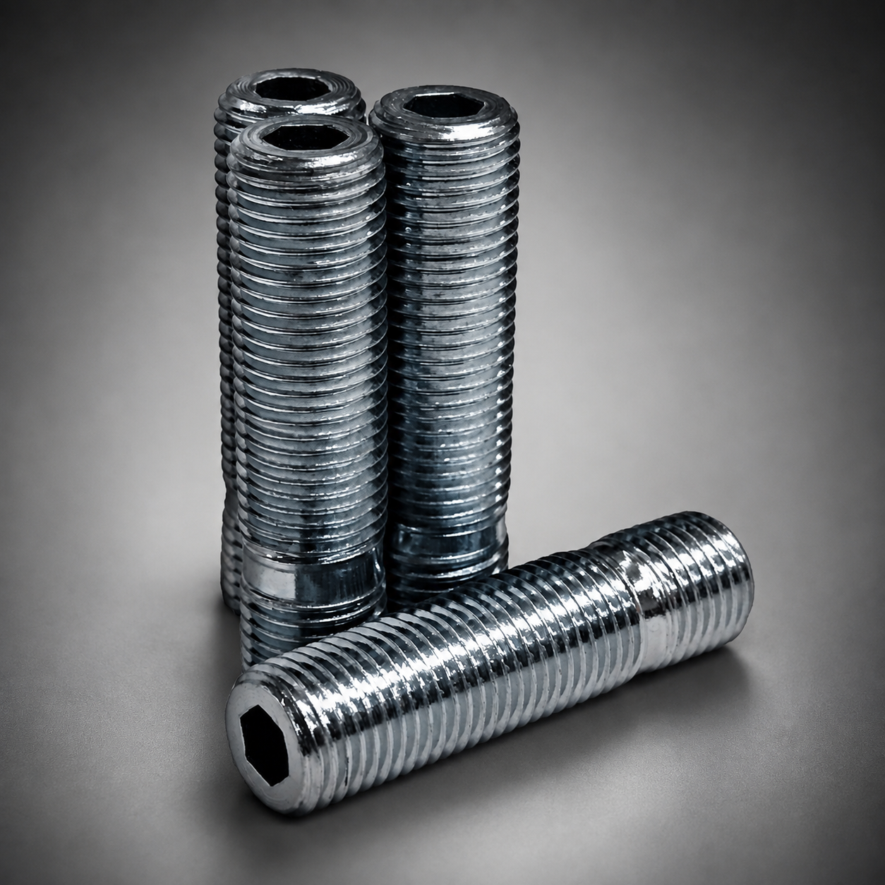 Four wheel stud conversion studs with threaded shanks, arranged with three standing vertically and one lying horizontally in the foreground on a smooth gray gradient background.
